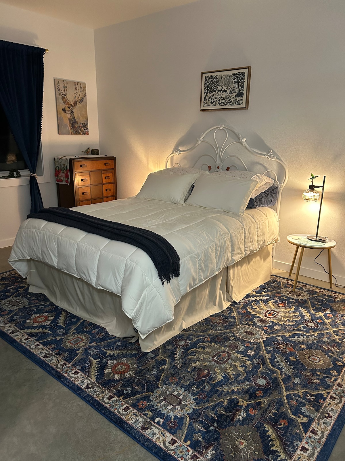 A spacious sleeping area is highlighted by a white bed with a light-colored quilt and decorative pillows. A stylish bedside table with a lamp and a patterned area rug add warmth. A chest of drawers and artwork adorn the walls, while dark curtains frame the window.