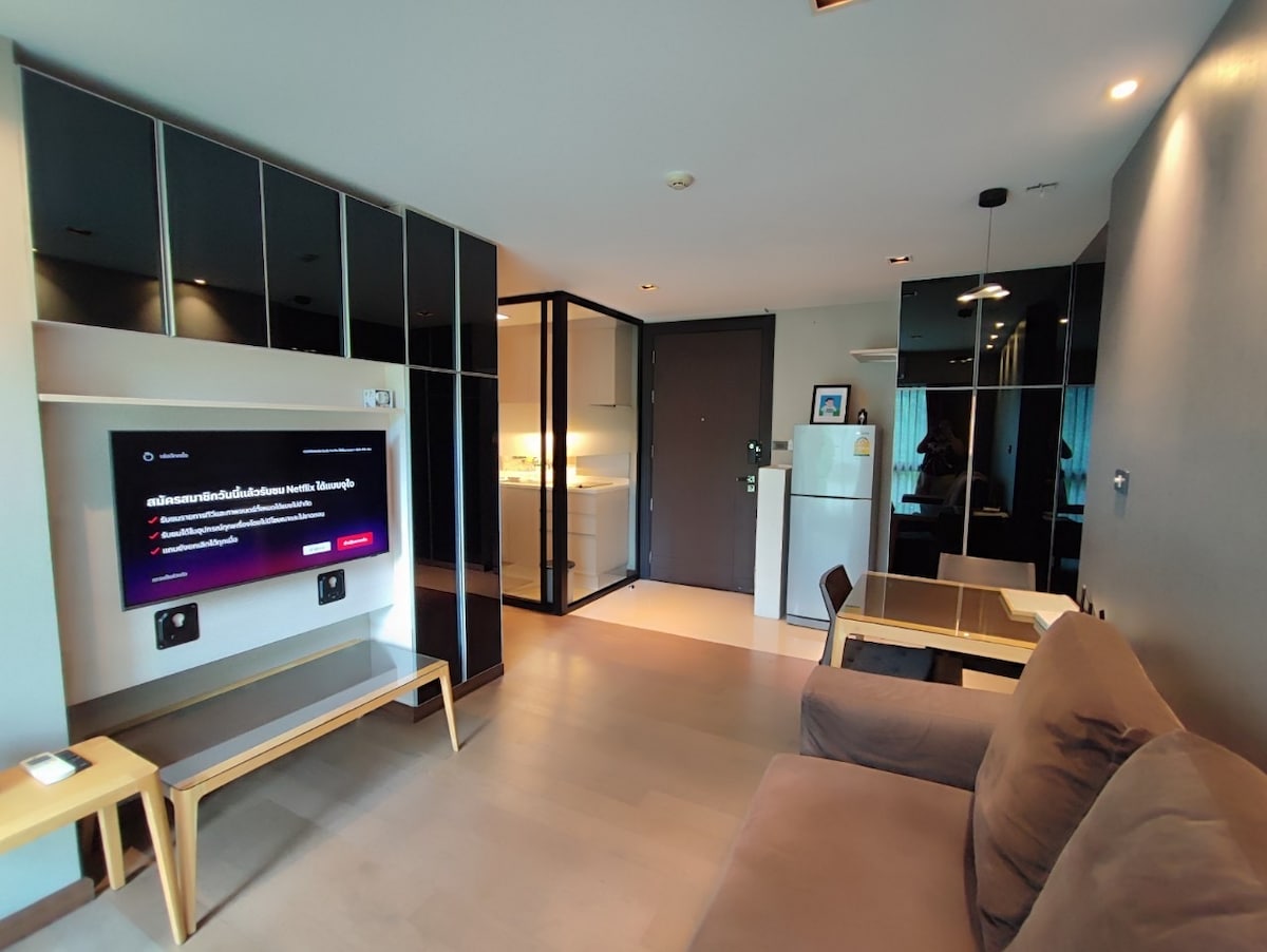 A modern living area is depicted, featuring a large flat-screen television mounted on a wall opposite a comfortable sofa. A glass partition leads to a bright kitchenette, equipped with a refrigerator and kitchen essentials. Neutral tones and minimalistic furniture create a stylish atmosphere.