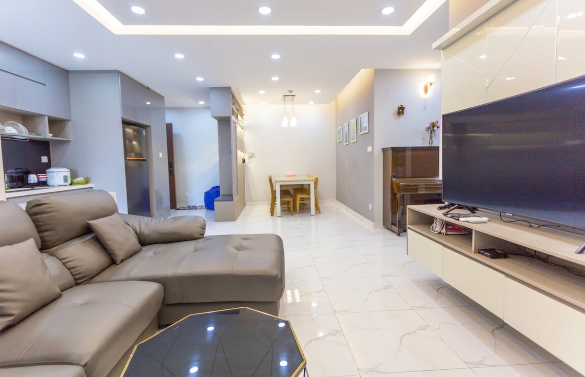 A spacious living area features a comfortable sectional sofa facing a large television. A dining table is positioned nearby, accommodating several people. The modern kitchen is visible in the background, and soft lighting enhances the open and airy feel of the space.