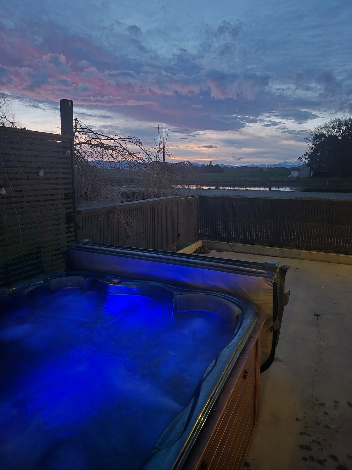 A spa pool is viewed in the foreground, displaying soft blue lighting that contrasts with the twilight sky. Beyond the pool, a serene landscape features a body of water and distant hills, with a wooden fence demarcating the outdoor space.