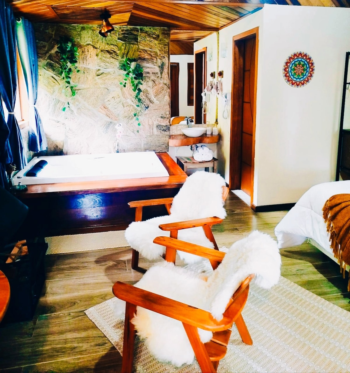 A warm interior space features a cozy ambiance with wooden accents and soft lighting. Two plush armchairs are positioned near a spacious bed, and a hot tub is visible in the corner, making the area inviting. Natural stone elements enhance the charm of the room.