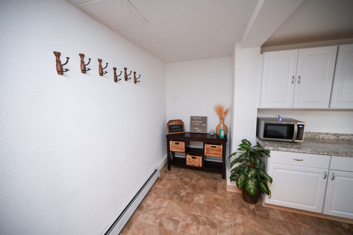 A spacious corner of the kitchen area features a wall with multiple hooks, a small shelf with woven baskets and a decorative plant. A microwave sits on the counter, surrounded by light-colored cabinets and granite countertops.