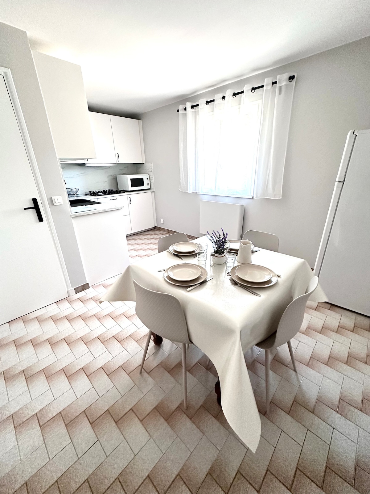 A bright kitchen area features a round table set for four, complete with plates and utensils. White cabinetry and a modern refrigerator are visible, along with a window that allows natural light to illuminate the space. The floor is designed with a herringbone pattern.