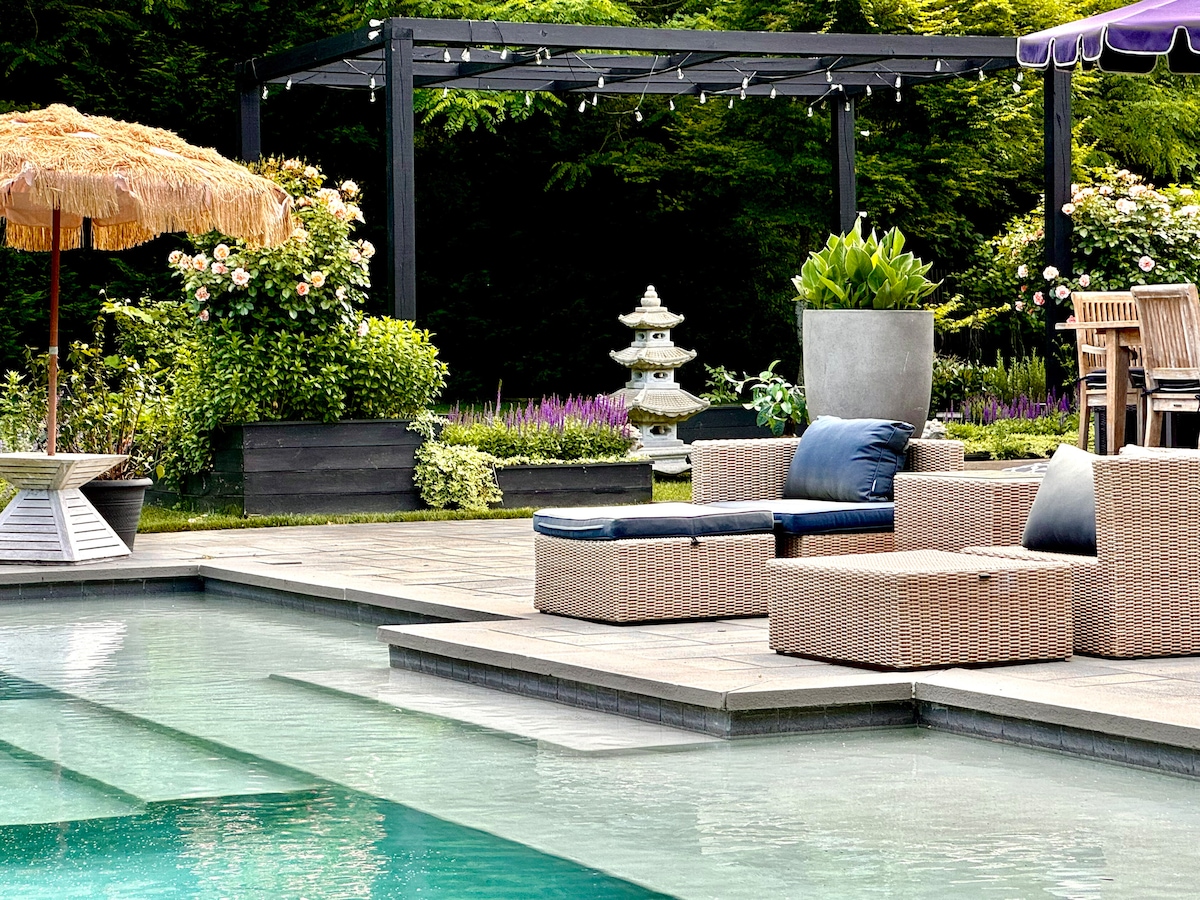 A tranquil outdoor space features a clear swimming pool with lounge chairs positioned nearby. A shaded seating area under a wooden pergola showcases comfortable seating and potted plants, while colorful flowers add visual interest to the landscape.