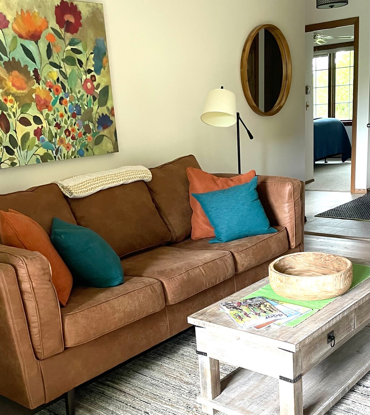 A comfortable living area features a brown sofa adorned with colorful cushions in shades of orange and teal. A wooden coffee table complements the space, holding a decorative bowl and magazines. A vibrant floral artwork is displayed on the wall, providing a cheerful focal point.