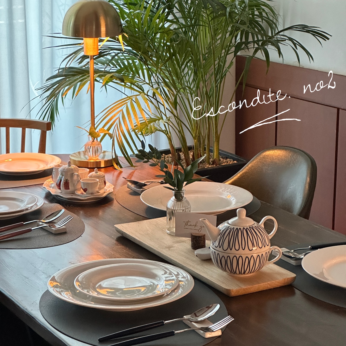An elegantly set dining table features white plates, cutlery, and a decorative teapot at the center. A small plant and a stylish lamp add warmth to the setting, while a wooden serving board holds dark chocolates. Natural light filters through sheer curtains.