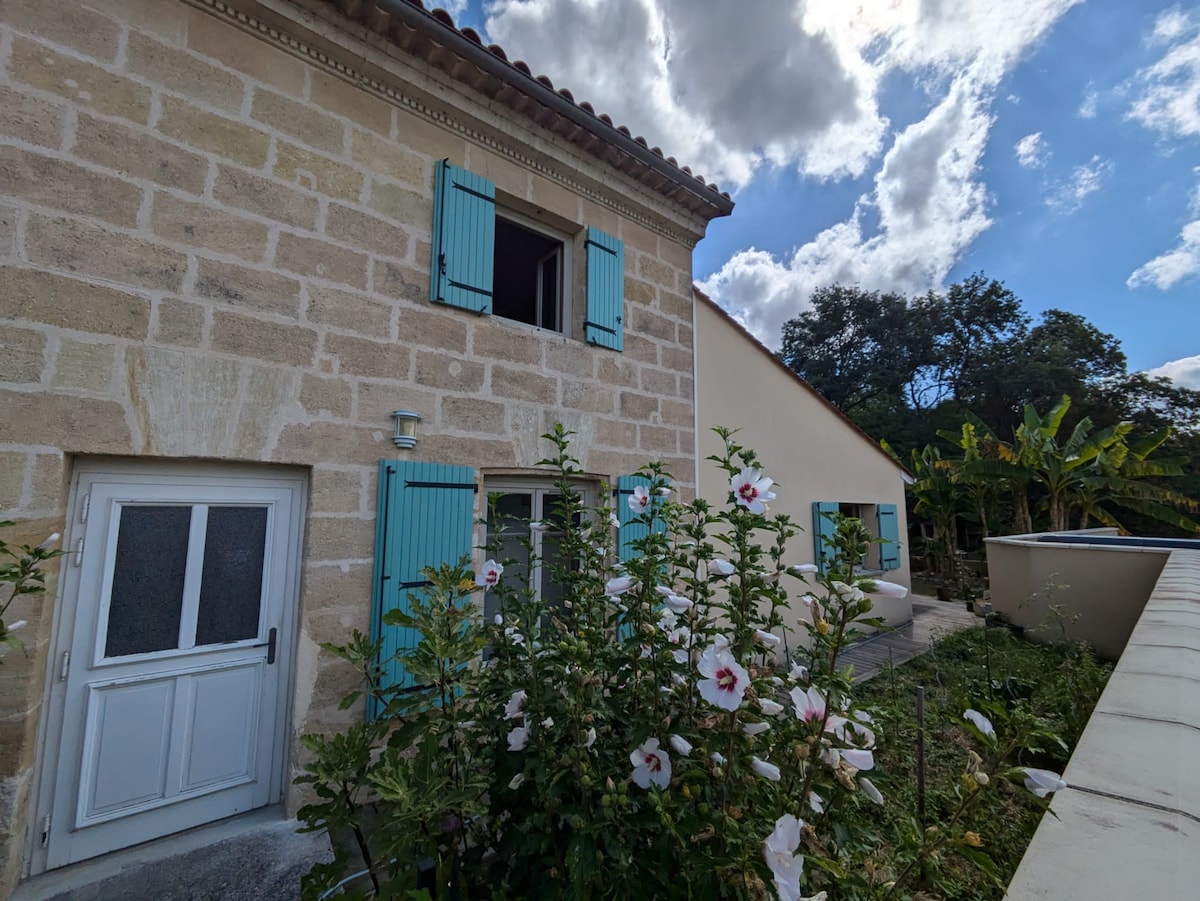 The exterior of a traditional stone house is displayed, featuring light blue shutters on the upper windows. A white door is visible, framed by lush greenery and blooming hibiscus flowers. The sky is partly cloudy, adding a serene atmosphere to the scene.