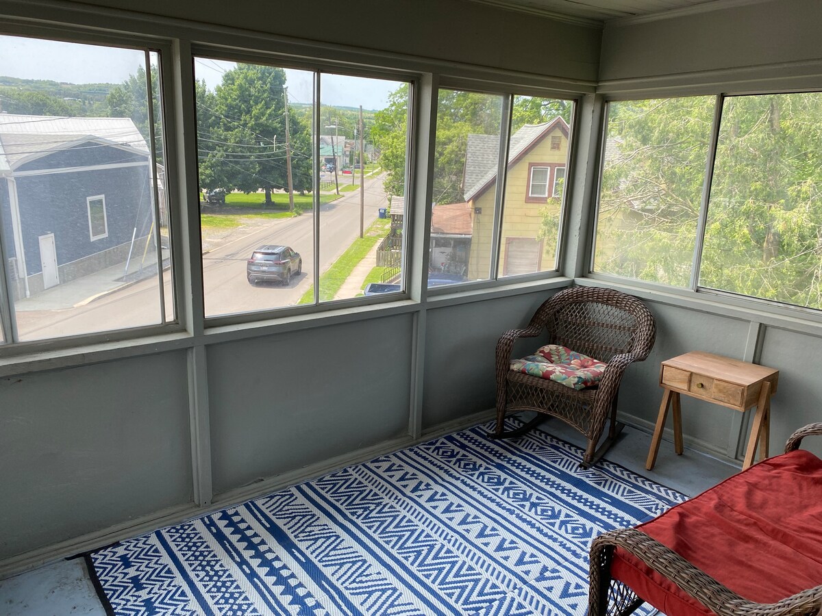 A screened-in porch is presented with large windows offering views of the street and nearby structures. A wicker chair with a floral cushion and a small side table are positioned on a patterned blue and white rug, allowing for relaxation in a breezy setting.