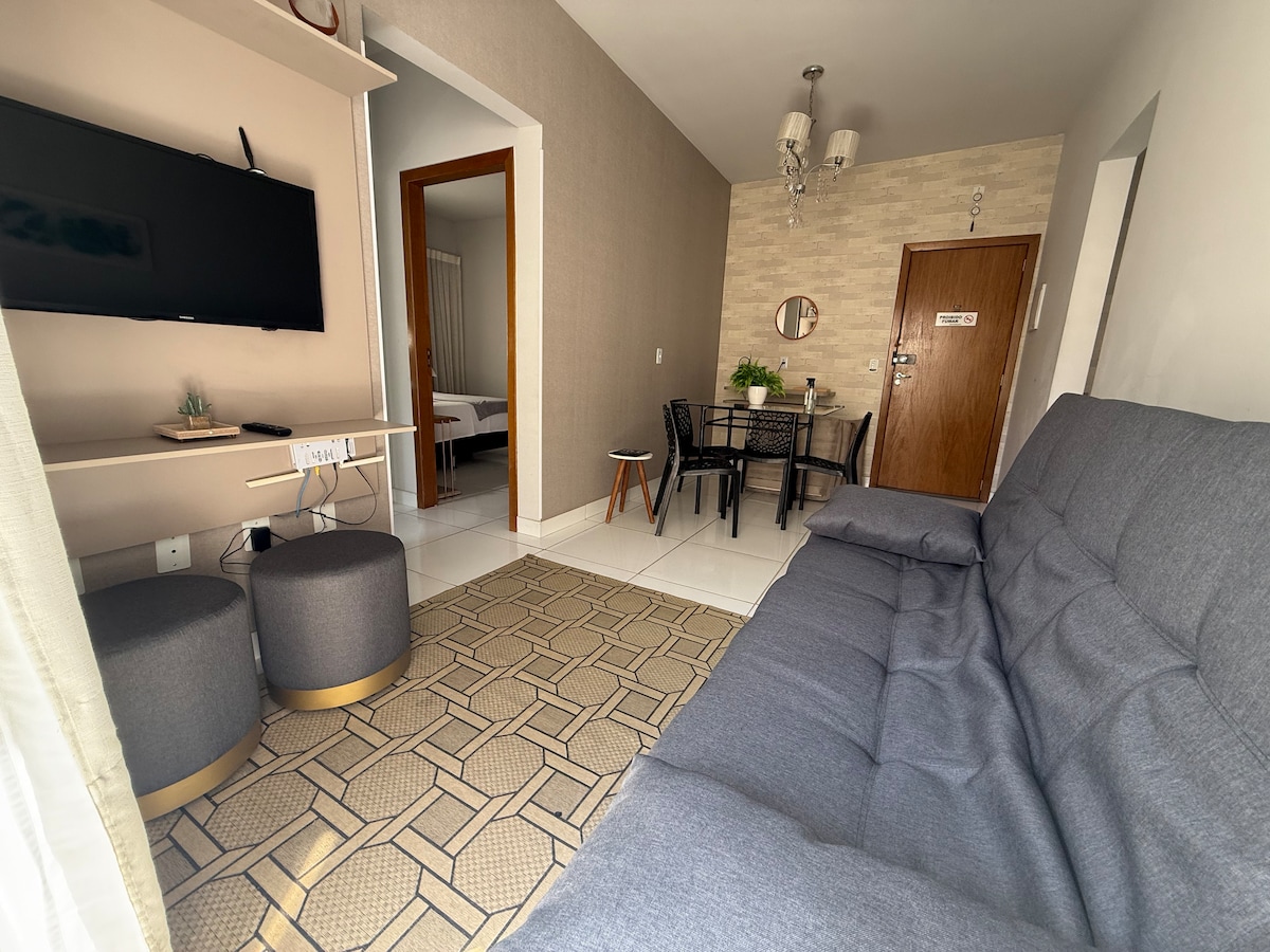 A neatly arranged living area features a gray sofa bed and two round ottomans. A wall-mounted television is positioned above a low shelf. A small dining table with chairs is visible in the background, along with a door leading to another room.