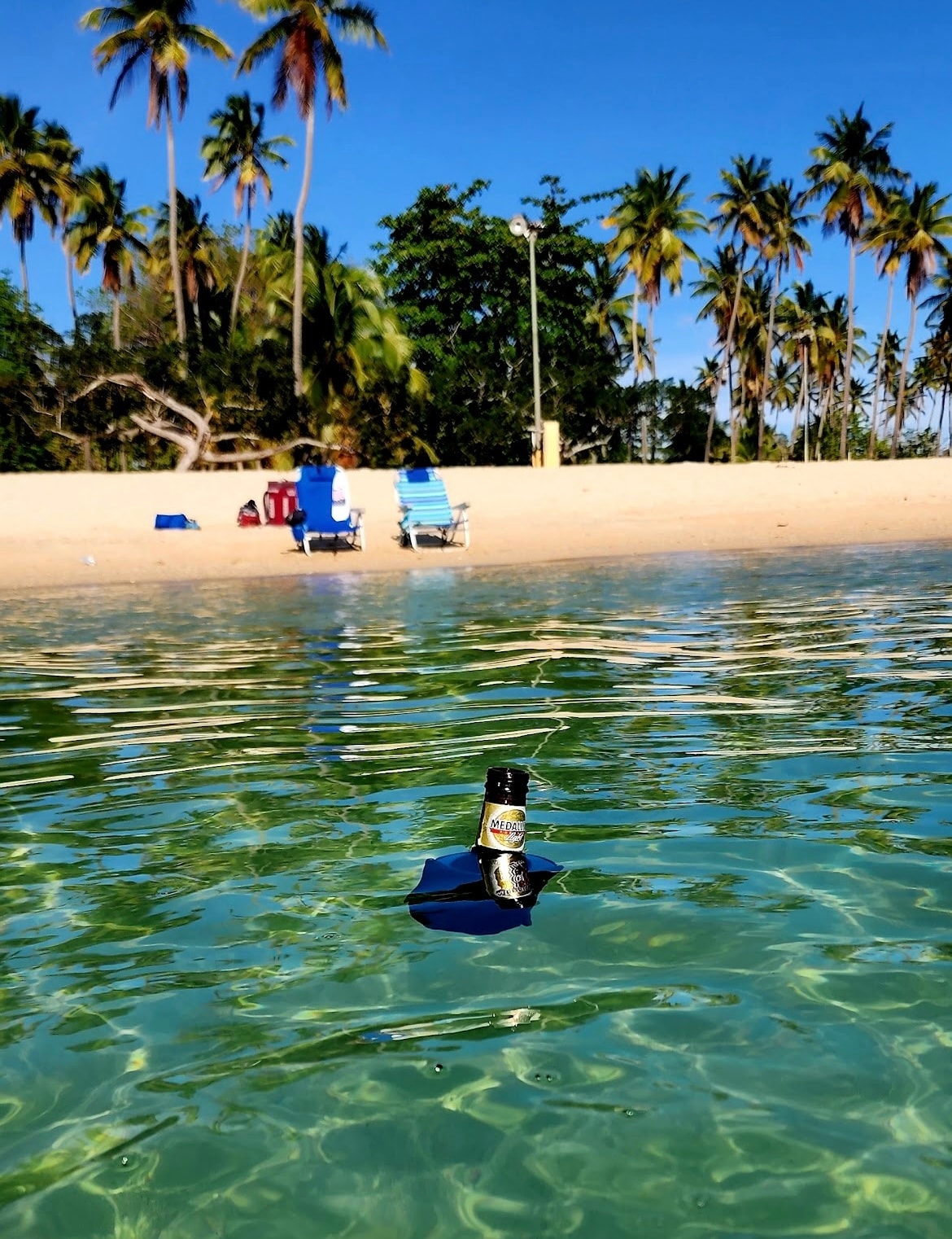 In the foreground, a bottle floats on clear water with rippling reflections. A sandy beach is visible in the background, framed by tall palm trees. Two beach chairs are positioned further back, creating a serene coastal vibe.