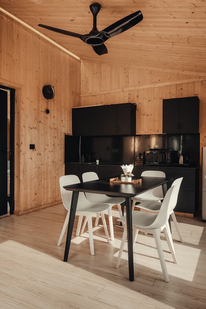 Një hapësirë ngrënieje e ngrohtë, e veshur me dru, me një tavolinë të zezë elegante dhe karrige moderne të bardha, e përsosur për vakte të përbashkëta, lojëra tavoline ose punë në distancë. Kuzhina e plotë prapa është e pajisur me gjërat thelbësore, duke e bërë të lehtë përgatitjen e çajit, kafesë ose ushqimeve të shpejta