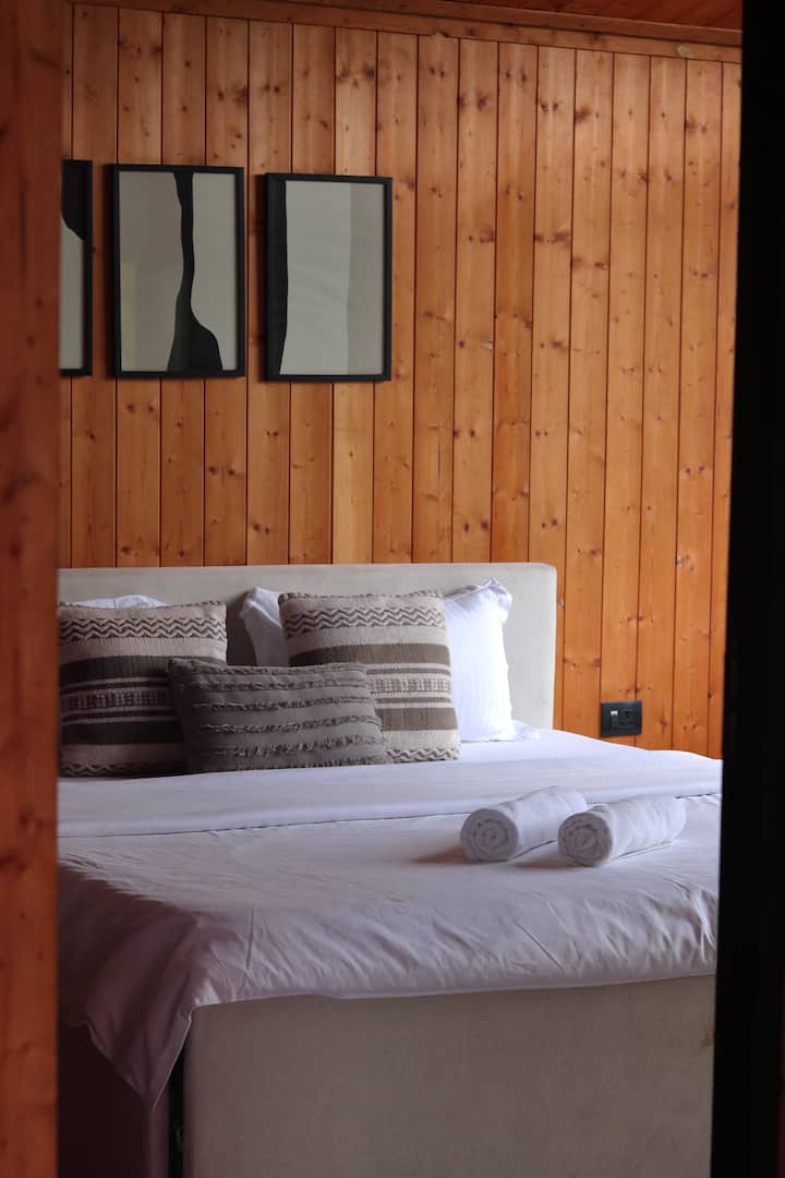 חדר שינה מרווח ומזמין עם מיטת קינג סייז, מצעים נקיים, מגבות מגולגלות ועיצוב פנים מעץ חם לשהייה רגועה ומנוחה.