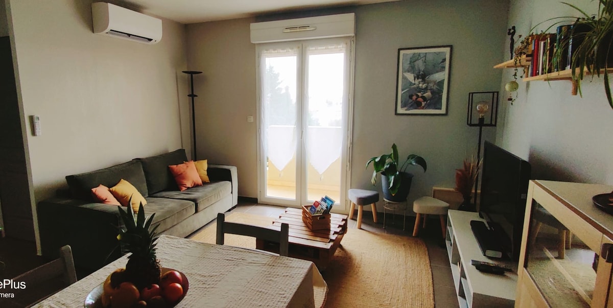 A cozy living area includes a comfortable sofa with vibrant cushions, alongside a low coffee table. Natural light floods the space through glass balcony doors, revealing a small terrace. A TV is positioned on a minimalist unit, with a plant and wall art adding a touch of warmth.