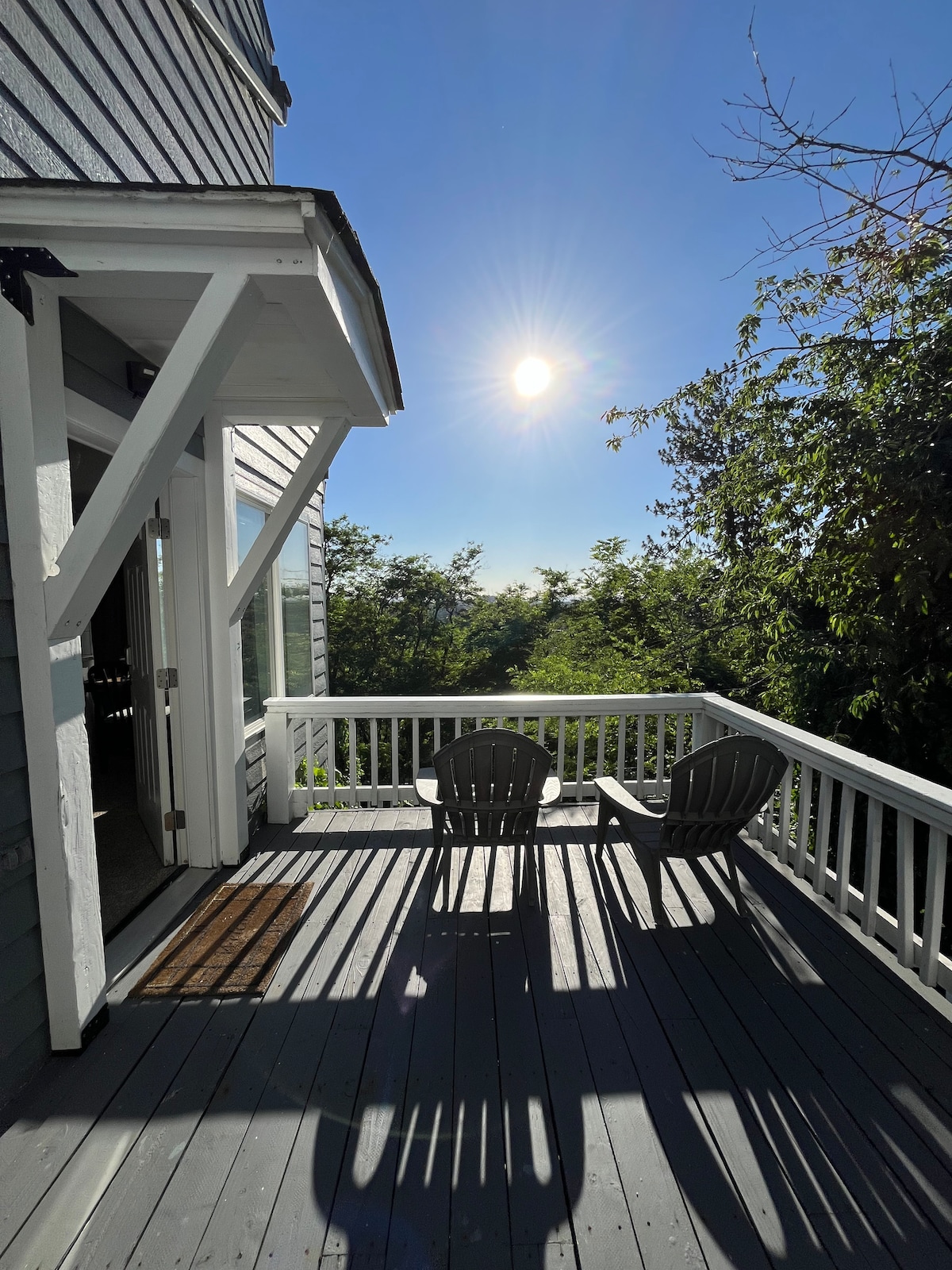 A private balcony is shown with two chairs positioned to take in the views. Sunlight filters through the surrounding greenery, creating a warm ambiance. The entrance to the studio is visible, inviting guests to enjoy the outdoor space.