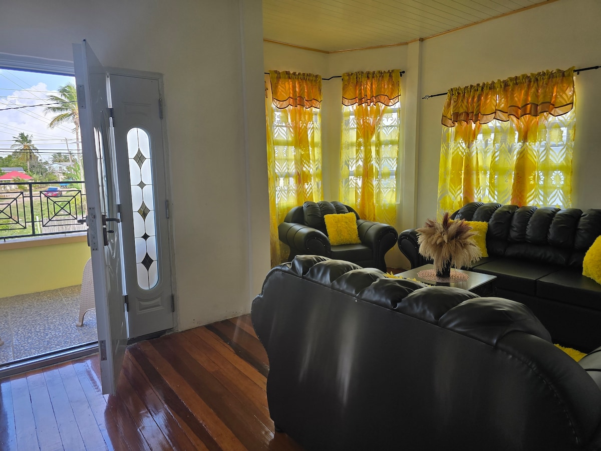 A bright living area features a comfortable black leather sectional sofa facing a stylish coffee table. Natural light filters through large windows adorned with cheerful yellow curtains, creating a warm atmosphere. A decorative plant adds a touch of greenery next to the entrance.