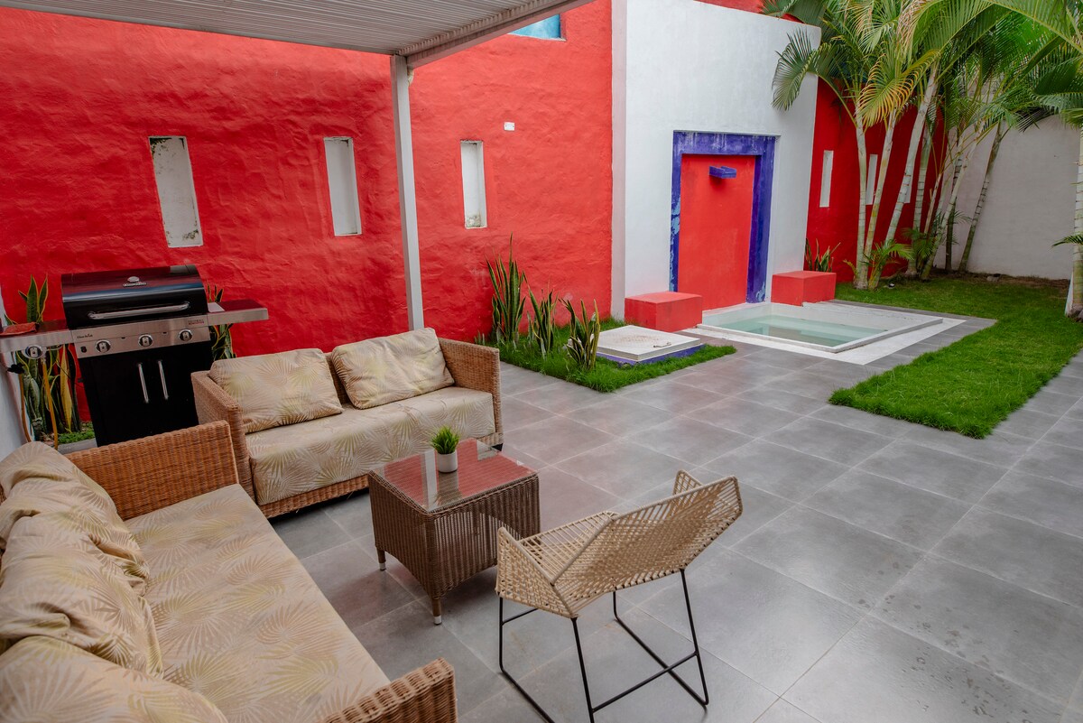 An outdoor patio area is framed by vibrant red walls, featuring comfortable seating and a small table. A barbecue grill is positioned nearby, alongside a manicured lawn and a tranquil water feature that adds a serene touch to the space.