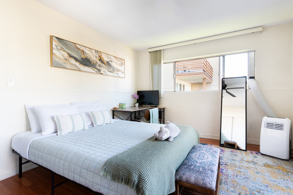 A bright and airy bedroom features a spacious bed dressed in a soft quilt, complemented by decorative pillows. Natural light filters through a large window, highlighting a cozy seating area with a bench. A mirror and small desk are positioned nearby.