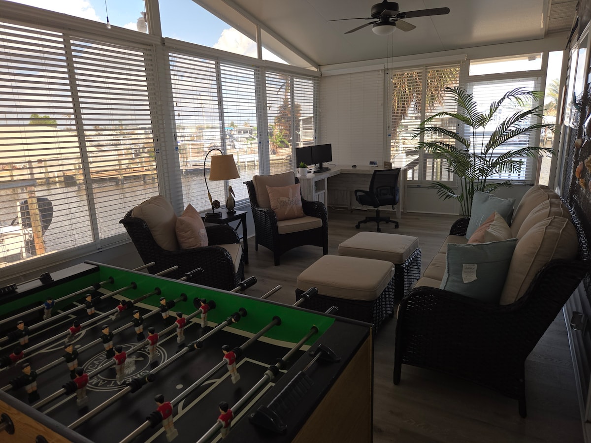 The enclosed lanai features a foosball table in the foreground, with a seating area that includes two wicker chairs and an ottoman. A desk with a computer and office chair is seen in the background, along with a potted plant and large windows providing a view of the canal.