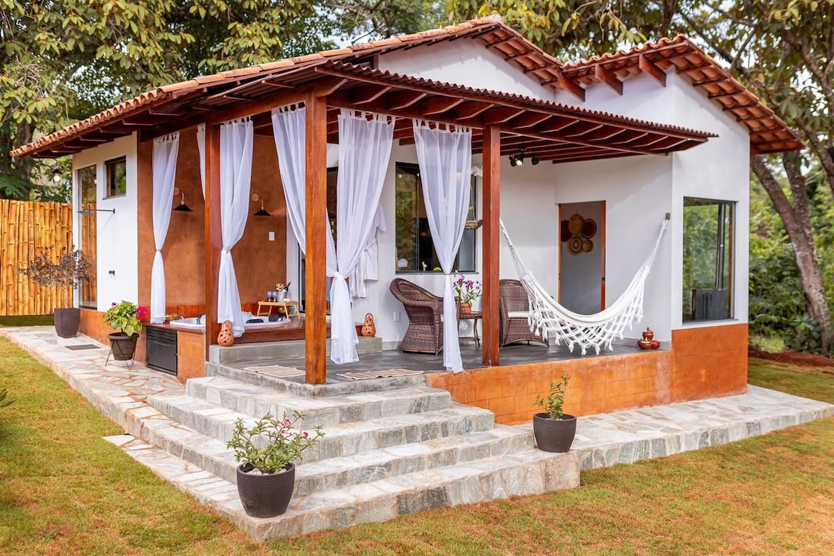 The exterior of a charming bungalow is depicted, featuring a stone staircase leading to a spacious patio. White drapes flow from the wooden posts, and a hammock is visible. Potted plants add greenery, enhancing the connection with the surrounding nature.
