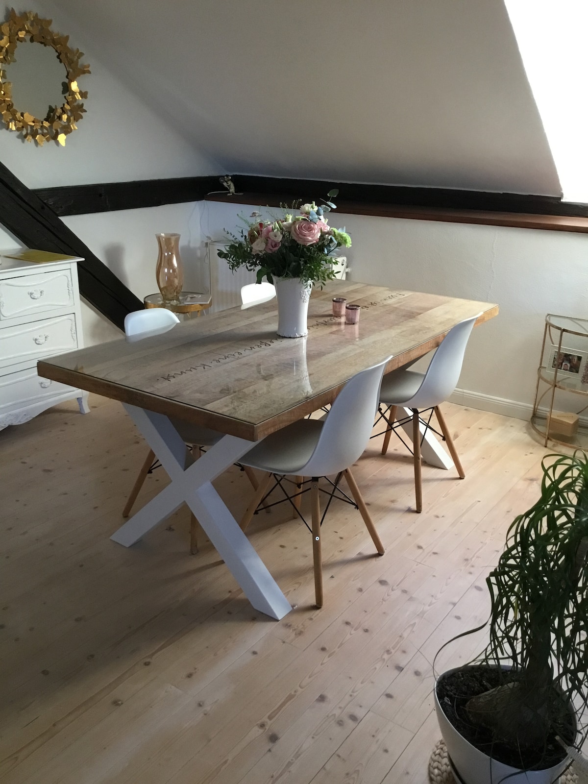A wooden dining table with a natural finish is surrounded by three white chairs featuring wooden legs. A vase with pink and white flowers sits at the center of the table, accompanied by two small candles. Light streams in from a nearby window, enhancing the inviting space.