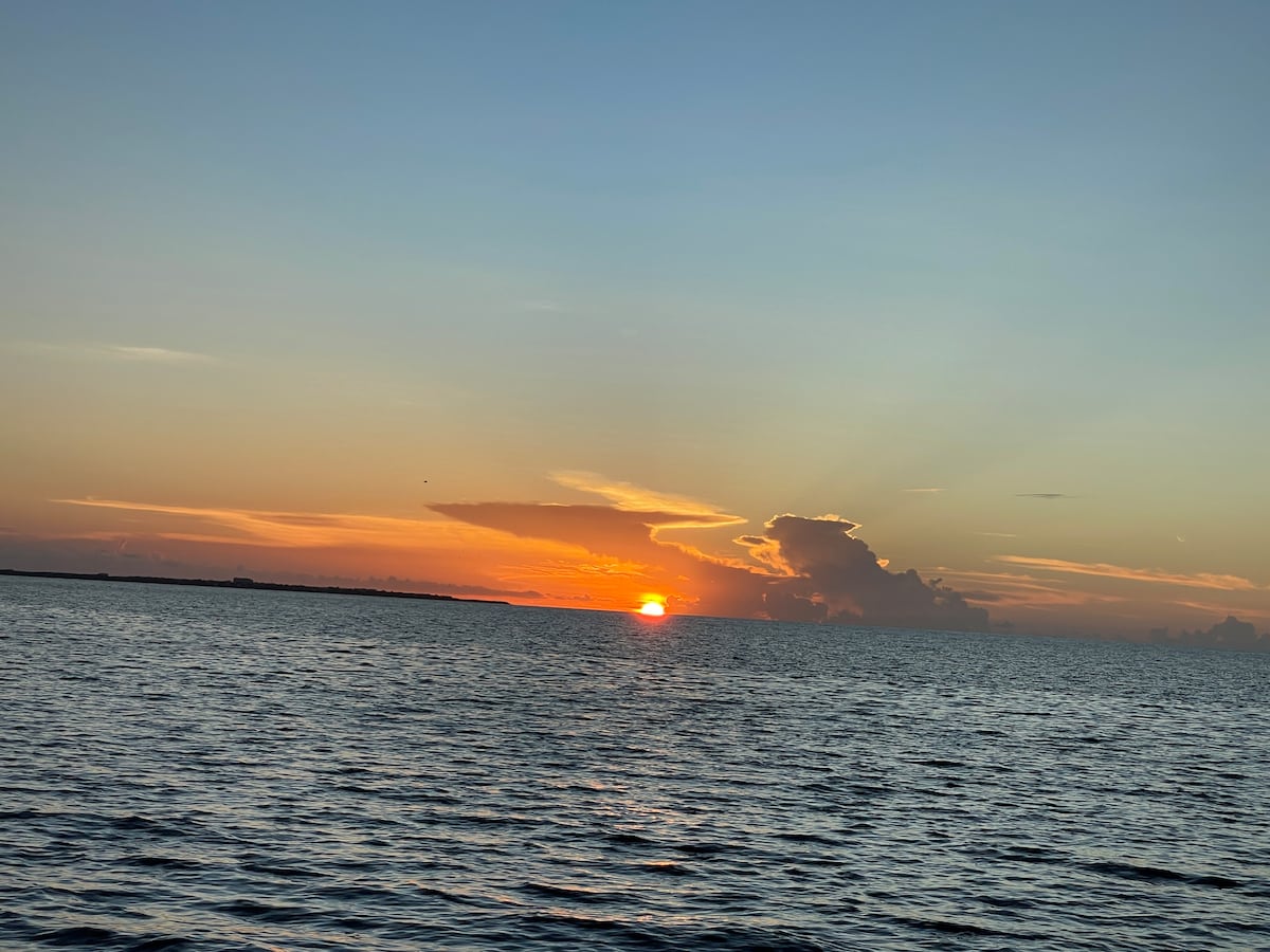 A serene view of the sun setting over the ocean, casting warm hues of orange and yellow across the sky. Gentle waves reflect the colorful sky, while clouds add dimension to the horizon. The tranquil water creates a peaceful atmosphere, enhancing the coastal experience.