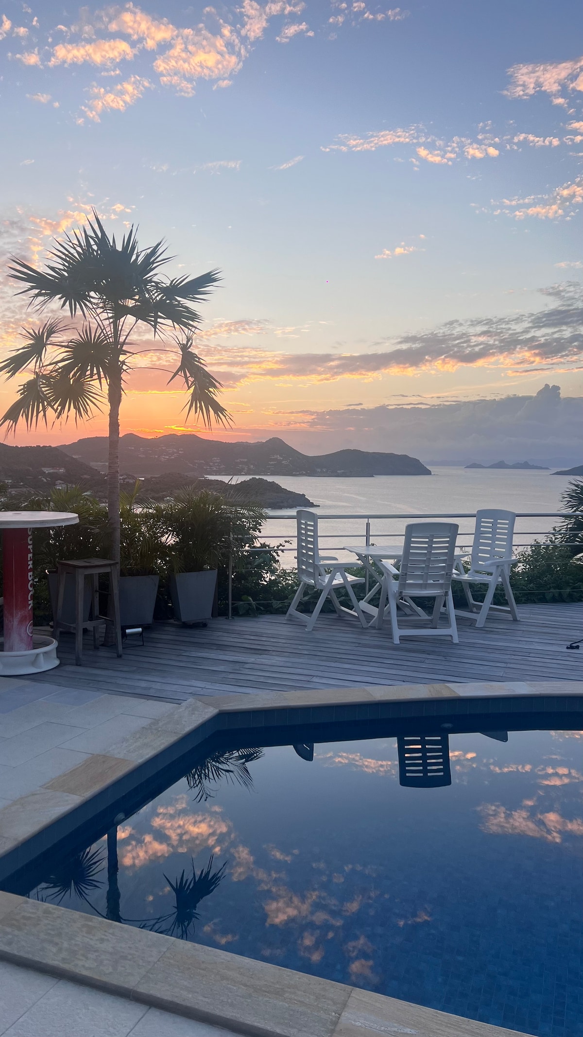 The image captures a serene sunset over the water, reflected in a private pool. A dining table with four white chairs is positioned near the pool, surrounded by tropical plants and hills that frame the scenic view.