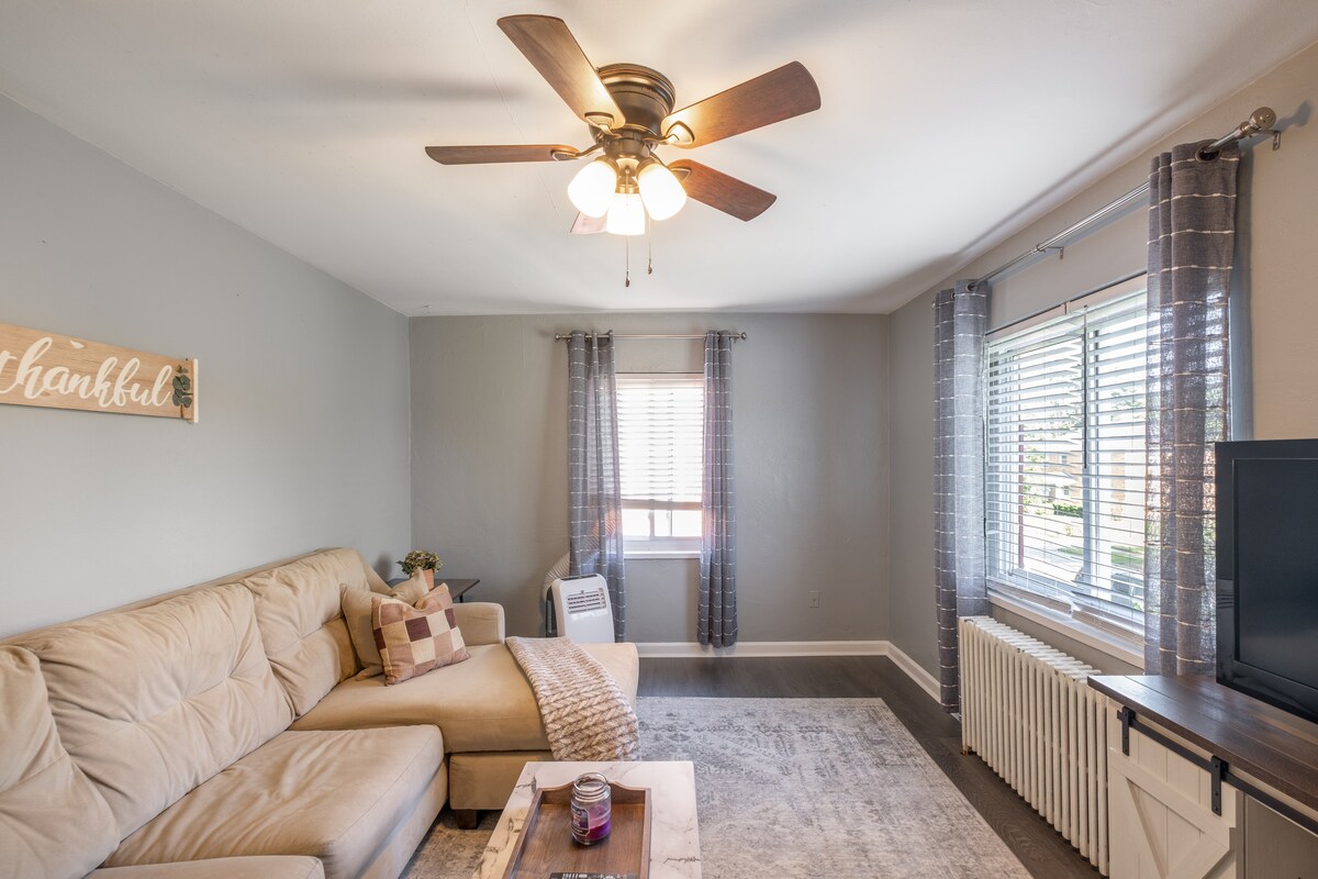 A spacious living room showcases a full-size sectional couch positioned against the wall. A coffee table sits in front, and a 50" Smart TV is in the corner. Natural light enters through the window, complemented by sheer curtains and a ceiling fan above.