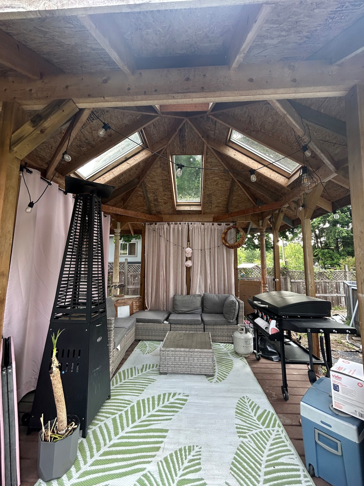 An open gazebo features a cozy seating area with a gray sectional sofa and a coffee table on a patterned rug. Natural light enters through skylights, complemented by soft curtains. A propane grill and cooler are positioned nearby, enhancing the outdoor entertainment space.