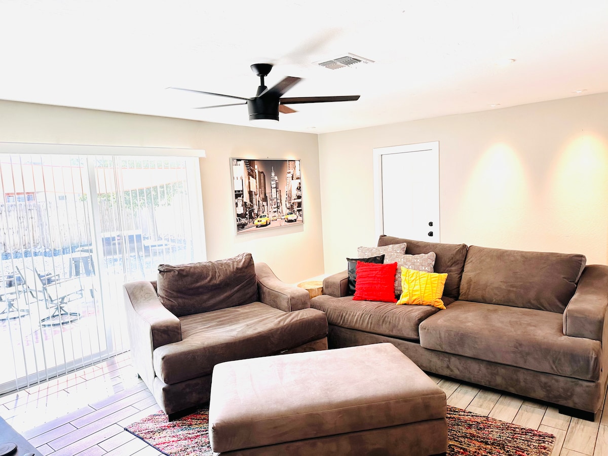 A spacious living area is furnished with a large sectional sofa, featuring colorful accent pillows. An ottoman sits in the center, while natural light pours in through sliding glass doors that lead to an outdoor space. A modern ceiling fan completes the room.