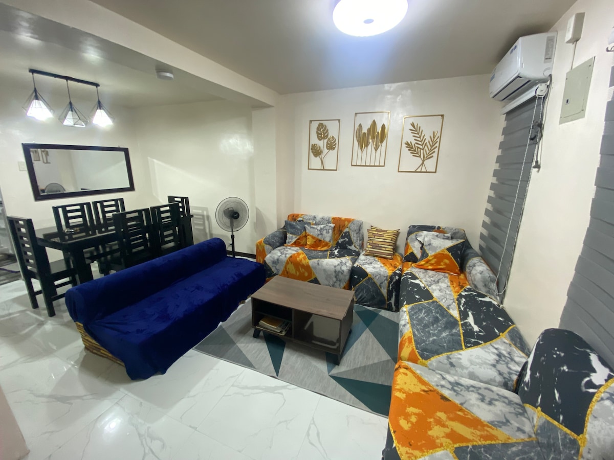 The spacious living area features a comfortable sofa set with a colorful geometric pattern, complemented by a wooden coffee table. A dining set for six is visible in the background, alongside a standing fan and modern light fixtures enhancing the room's ambiance.