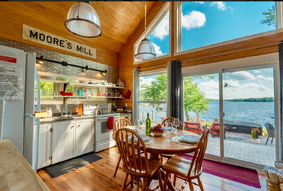 An inviting kitchen and dining area features wood accents and large windows overlooking the lake. A round wooden table is set for meals, accompanied by six chairs. Shelves hold various dishes and glassware, while a refrigerator stands nearby, completing the cozy atmosphere.