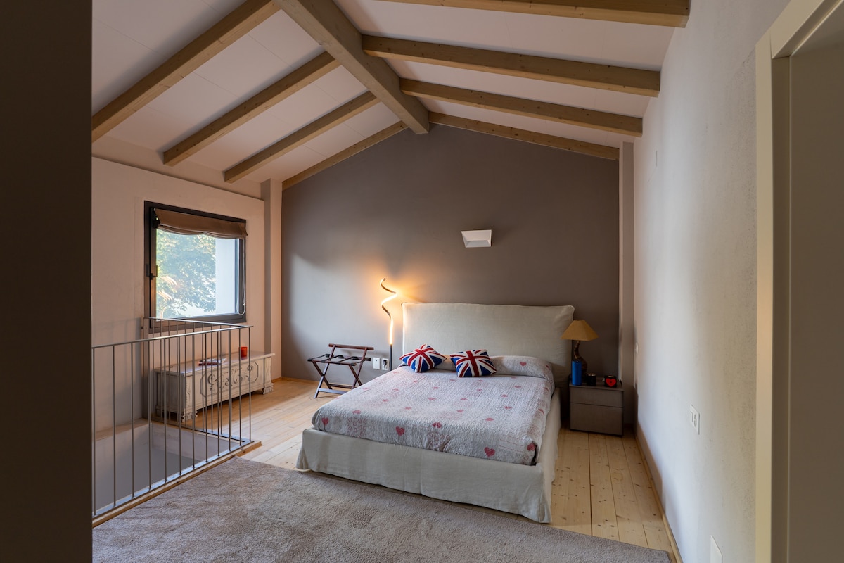 A spacious bedroom features a high ceiling with wooden beams. A large bed with a light cover is accented by decorative pillows. A small nightstand and a lamp are positioned beside the bed, while a seating area can be seen near the window, providing comfort and relaxation.