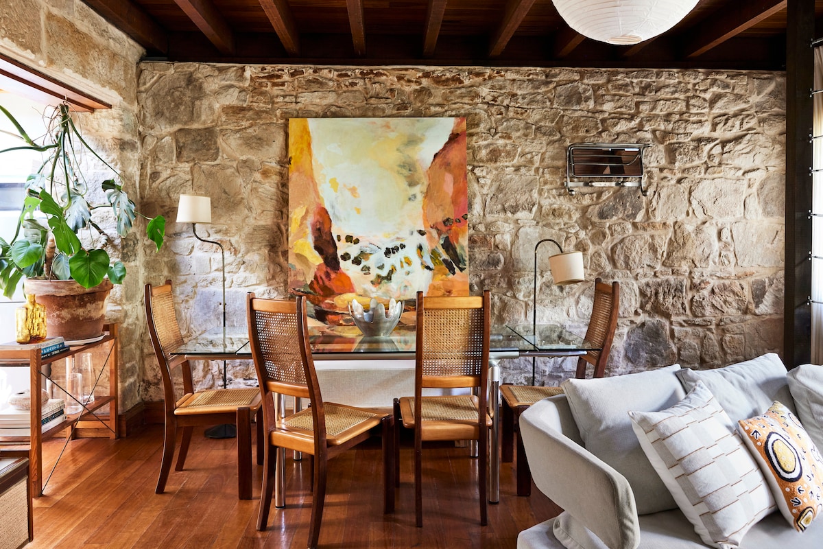 A dining area features a glass table surrounded by four wooden chairs. A large painting with abstract colors adorns the internal sandstone wall. A leafy plant is positioned near the window, while warm wooden flooring complements the inviting atmosphere.