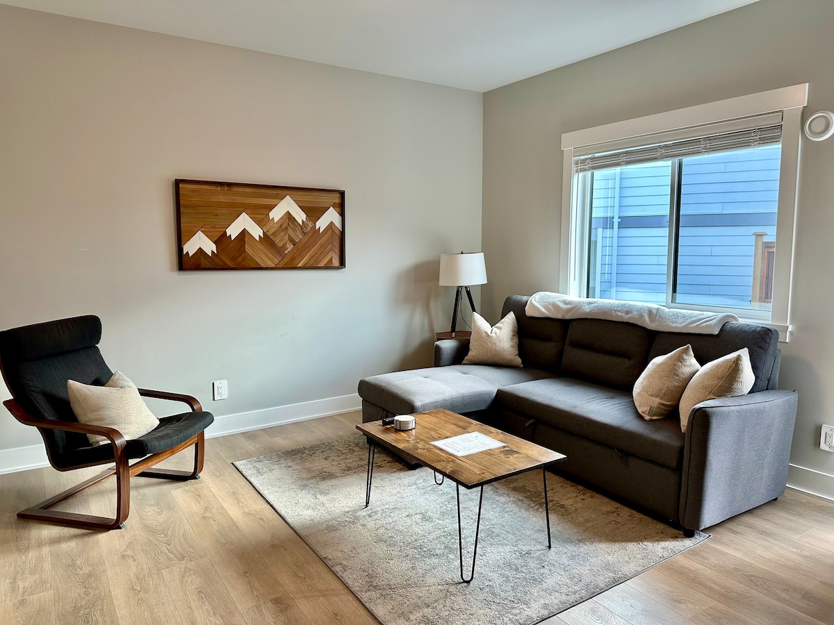 A living room is depicted, featuring a sectional sofa with decorative pillows, a wooden coffee table, and a stylish armchair. A lamp adds soft lighting, and a mountain-themed artwork hangs on the wall. Natural light enters through a window, enhancing the space.