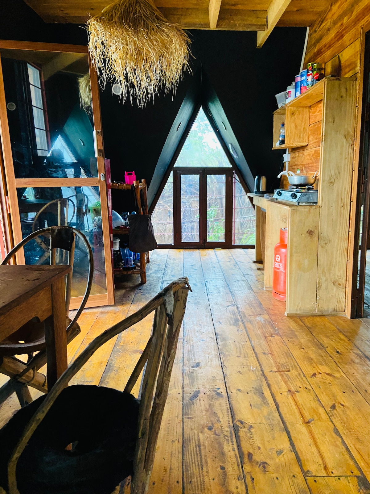 An interior view of the cabin showcases natural wooden flooring and sloped ceilings. A spacious kitchenette is visible, alongside a dining area with a wooden table and chairs. Large windows invite light and provide a view of the surrounding greenery.