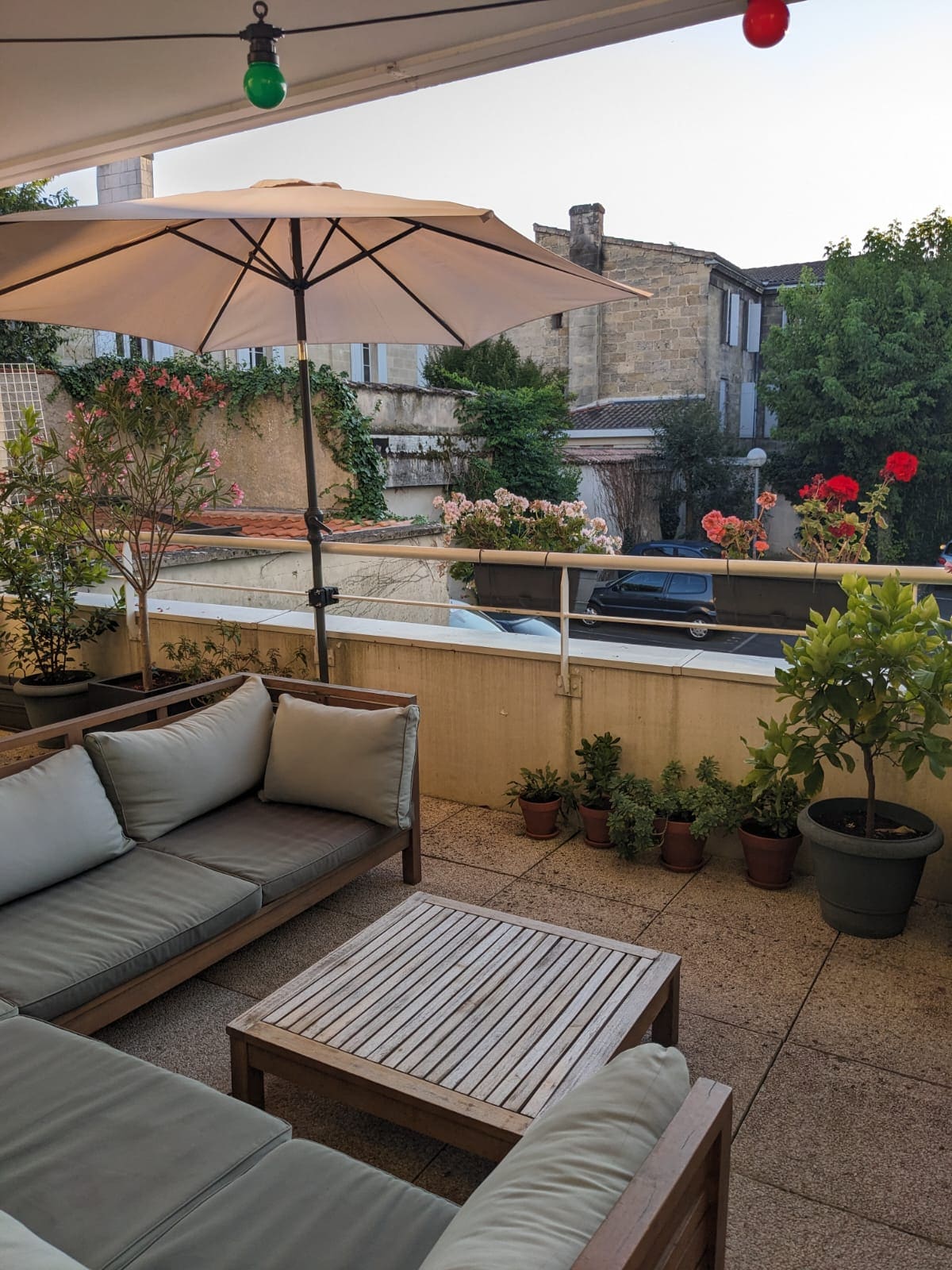 A spacious balcony is equipped with comfortable seating and a wooden coffee table, surrounded by vibrant plants and flowers. An umbrella provides shade, enhancing the outdoor space's accessibility and charm. The tranquil setting is complemented by a gentle view of nearby buildings and greenery.