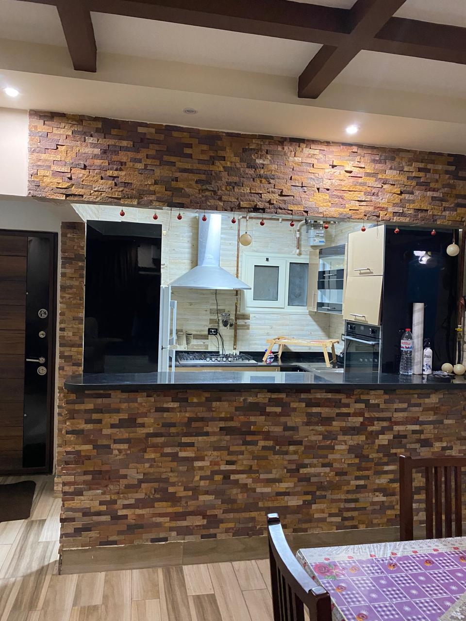 An open kitchen area is highlighted by a decorative stone wall and modern appliances. A black countertop separates the kitchen from a dining space, with soft lighting creating a welcoming ambiance. Wooden dining chairs are positioned around a table, enhancing the functional layout.