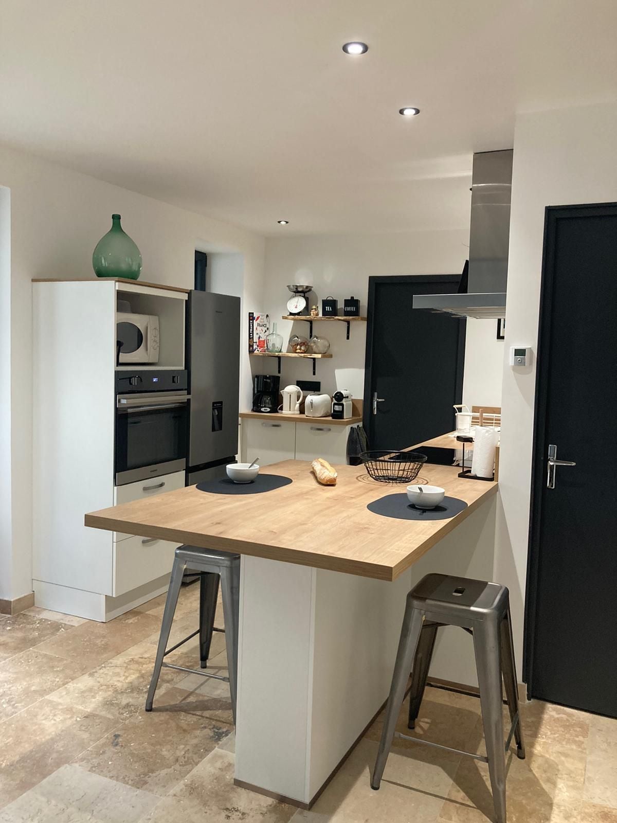 A modern kitchen is displayed, featuring sleek cabinetry and a central island with two metal stools. Essential appliances, including an oven, microwave, and coffee machine, are visible. Natural light brightens the space, highlighting the neutral-colored stone flooring.
