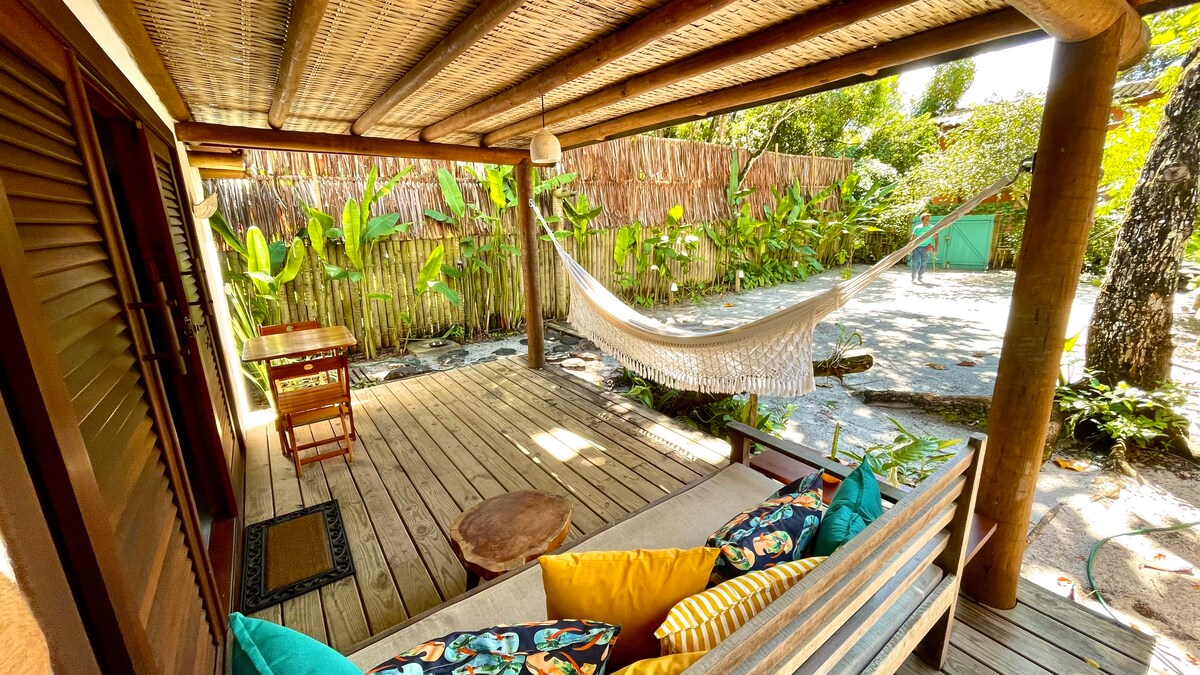 A shaded outdoor area features a wooden deck with a cozy seating arrangement and colorful cushions. A hammock is suspended between posts, inviting relaxation. Lush greenery surrounds the space, providing a natural and tranquil atmosphere.