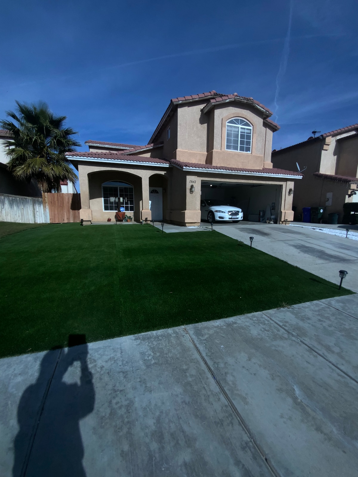 A two-story home is depicted with a well-maintained front yard featuring artificial grass. A driveway leads to a garage, with a vehicle parked inside. Palm trees add greenery to the exterior, and a clear blue sky complements the scene.