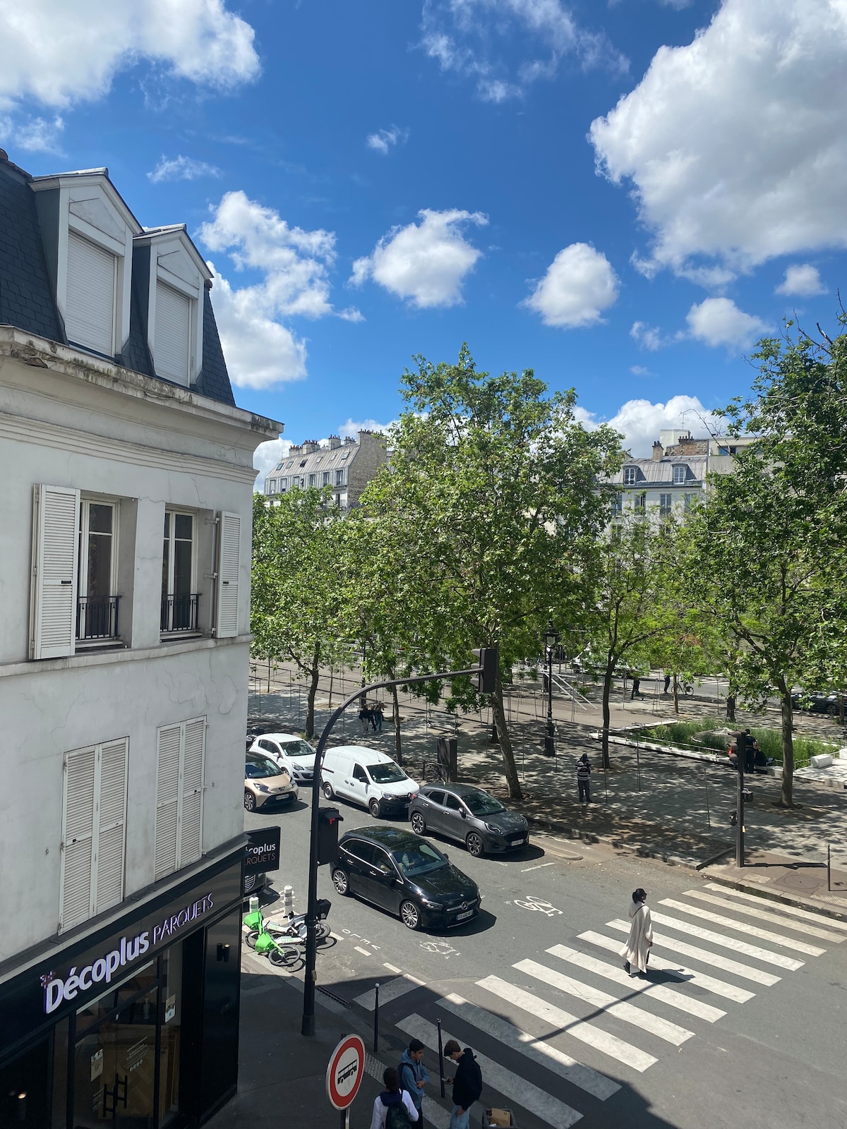 A view from a window overlooks a tree-lined boulevard. Vehicles are visible on the street, and pedestrians can be seen crossing at a marked intersection. The sky is dotted with fluffy clouds, adding a bright atmosphere to the scene.