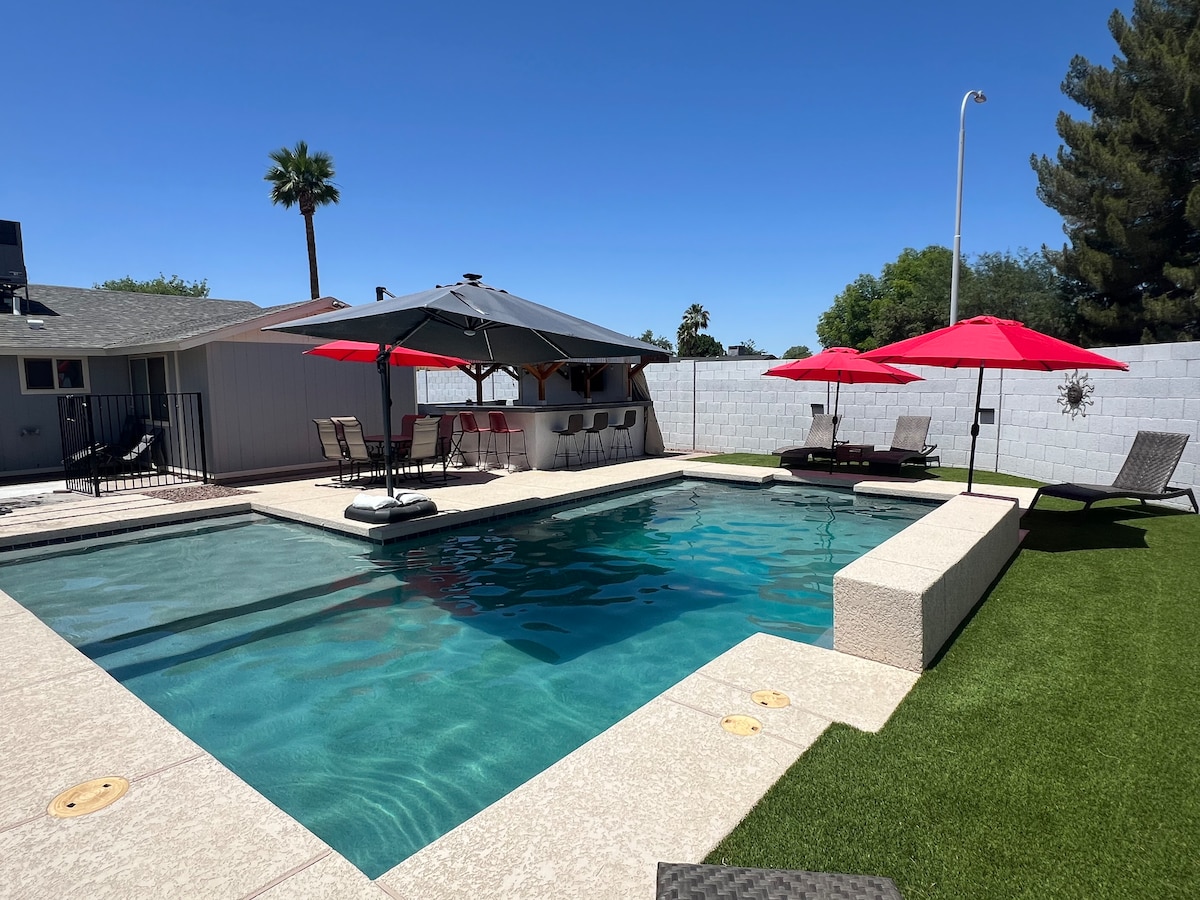 A spacious backyard features a climate-controlled pool surrounded by a paved area and artificial grass. Four red umbrellas provide shade over lounging chairs. A barbecue station is visible under a covered area with seating, ensuring a comfortable outdoor experience.