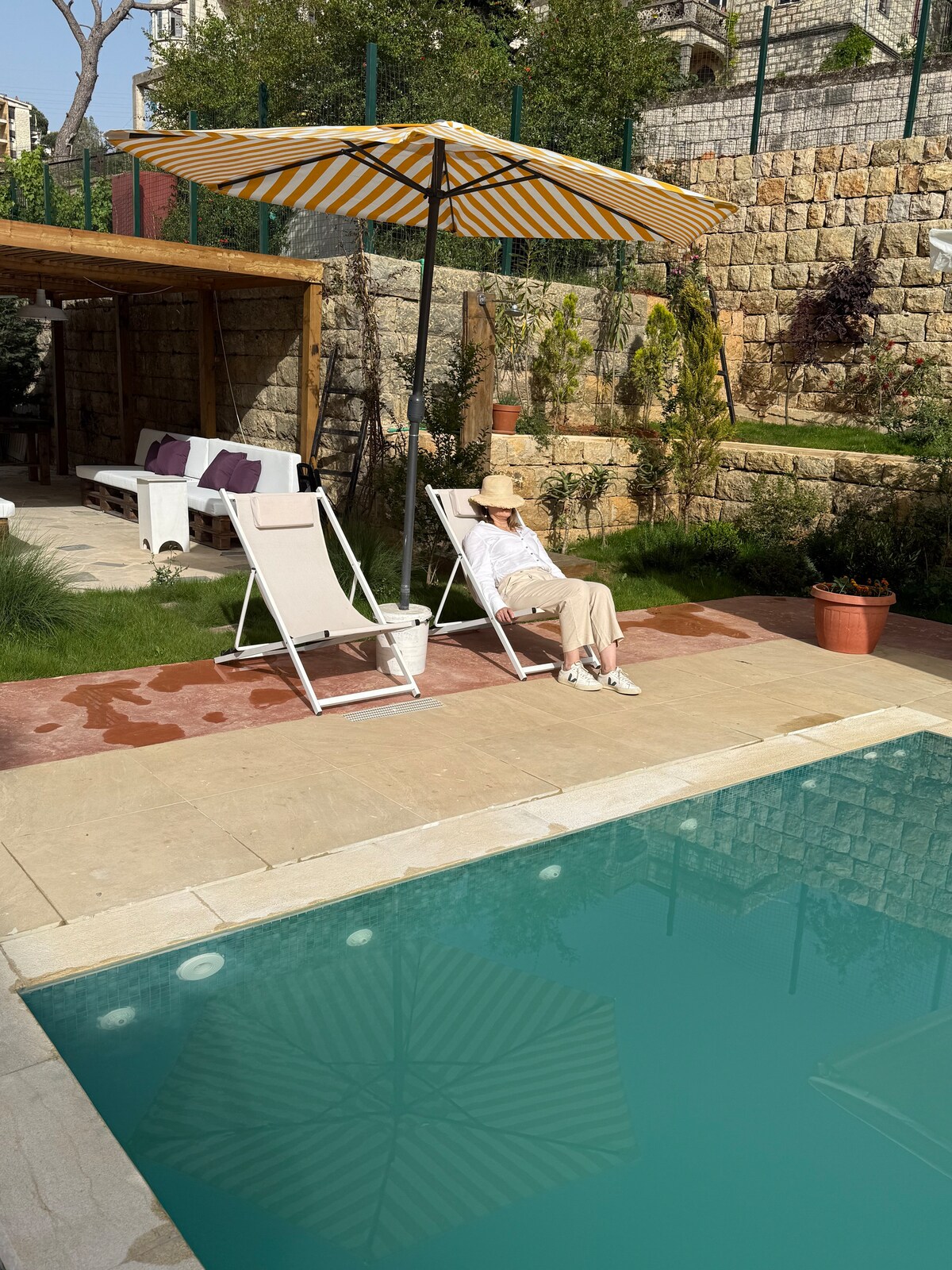 A relaxing pool area features a clear turquoise swimming pool surrounded by stone tiles. Two sun loungers are positioned beneath a striped umbrella, with a small potted plant nearby. Lush greenery and stone walls create a serene backdrop.
