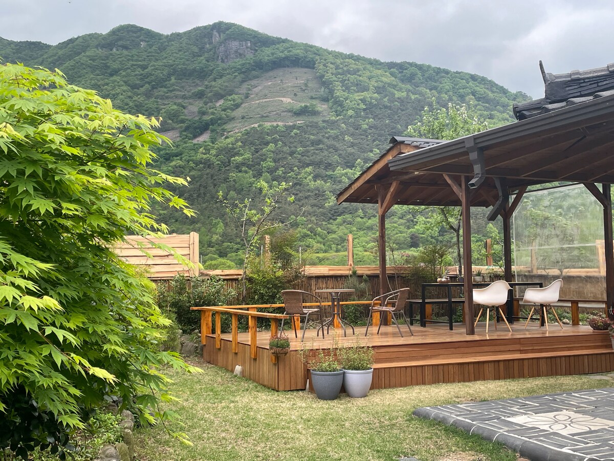 An outdoor deck surrounded by vibrant greenery offers a serene view of the mountains. The space is furnished with chairs and a table, providing an inviting area for relaxation or dining. Potted plants add a touch of color to the natural setting.