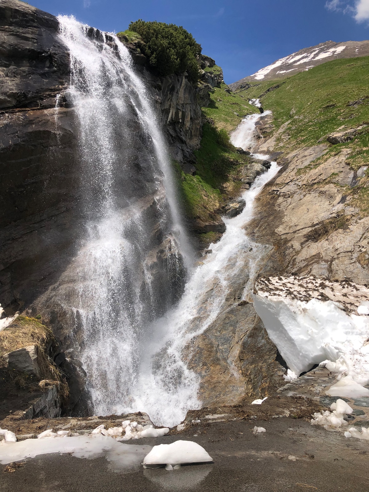 A cascading waterfall flows down rocky cliffs, surrounded by lush green vegetation. Ice and snow are visible at the base, contrasting with the water's movement. The backdrop features steep mountains under a blue sky, enhancing the natural scenery.