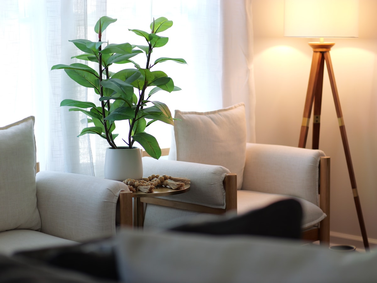 A cozy seating area features two light-colored armchairs with wooden frames. A potted plant adds a touch of greenery, and a small plate of snacks rests on a side table. Soft light emanates from a stylish floor lamp, enhancing the inviting atmosphere.