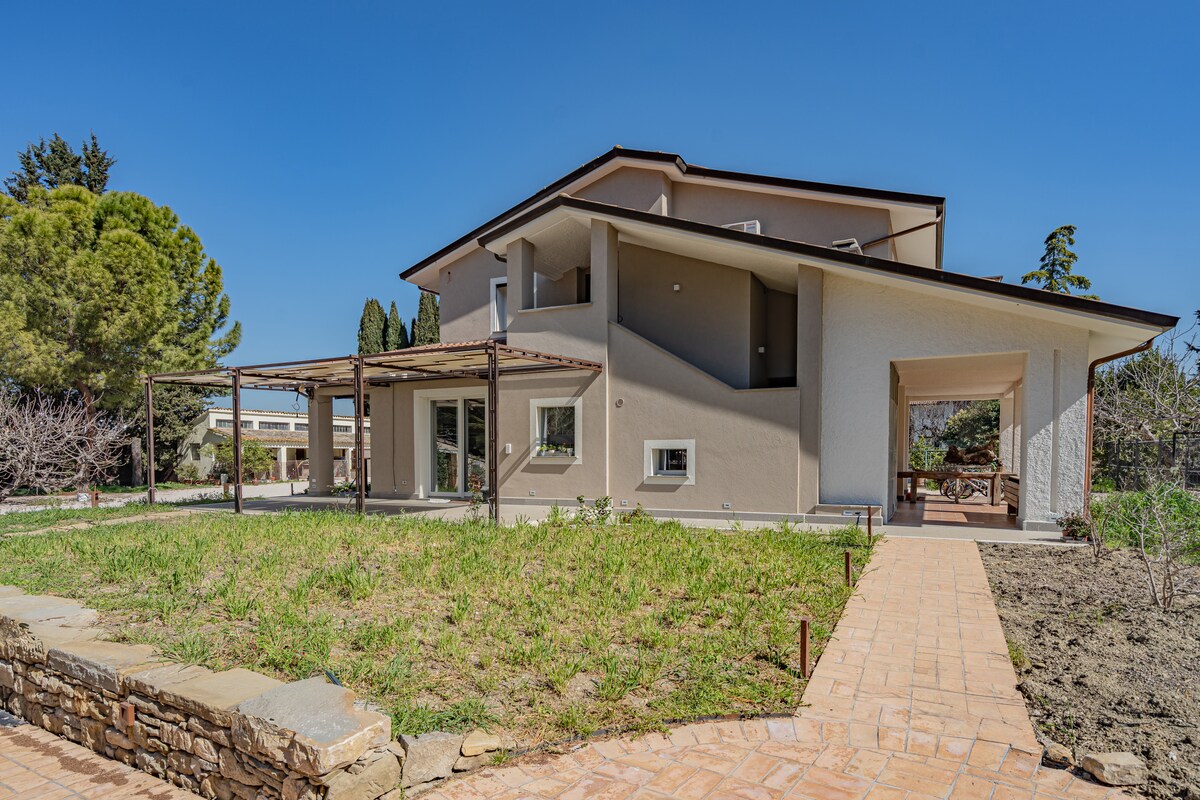 The exterior of a spacious house is surrounded by greenery and a manicured lawn. A stone pathway leads to a covered patio area with wooden beams, and a large patio opens to the garden, showcasing an inviting outdoor space for relaxation.