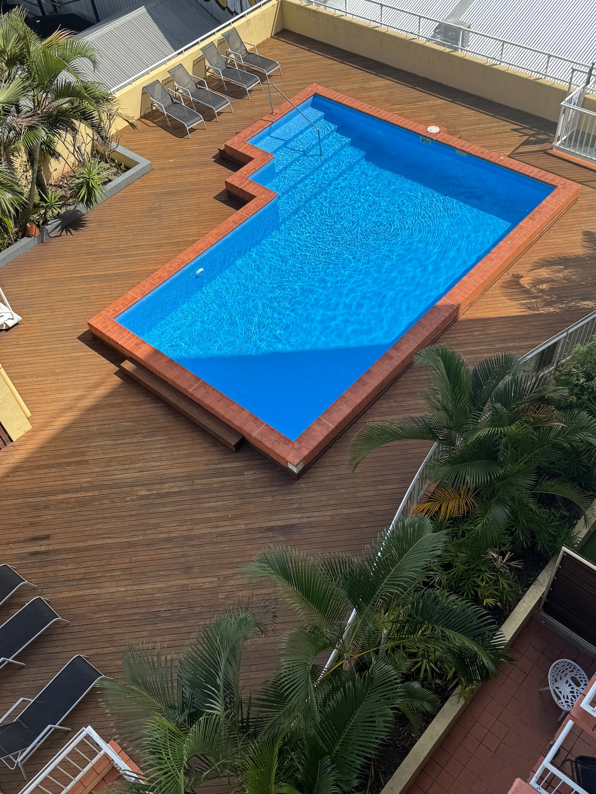 An outdoor pool designed in a unique shape is surrounded by wooden decking and lush greenery. Sun loungers are placed along the side of the pool, providing ample lounging space in a bright and inviting setting.