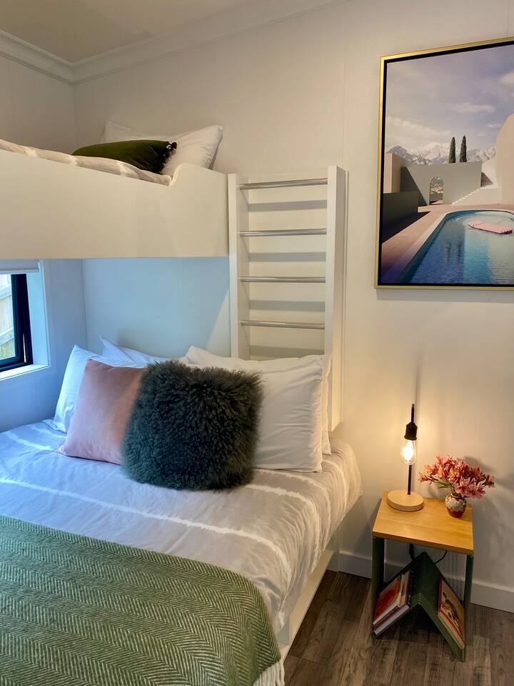 Modern bedroom with a white bunk bed, green and pink pillows, a cozy throw, and a nightstand with a lamp and flowers