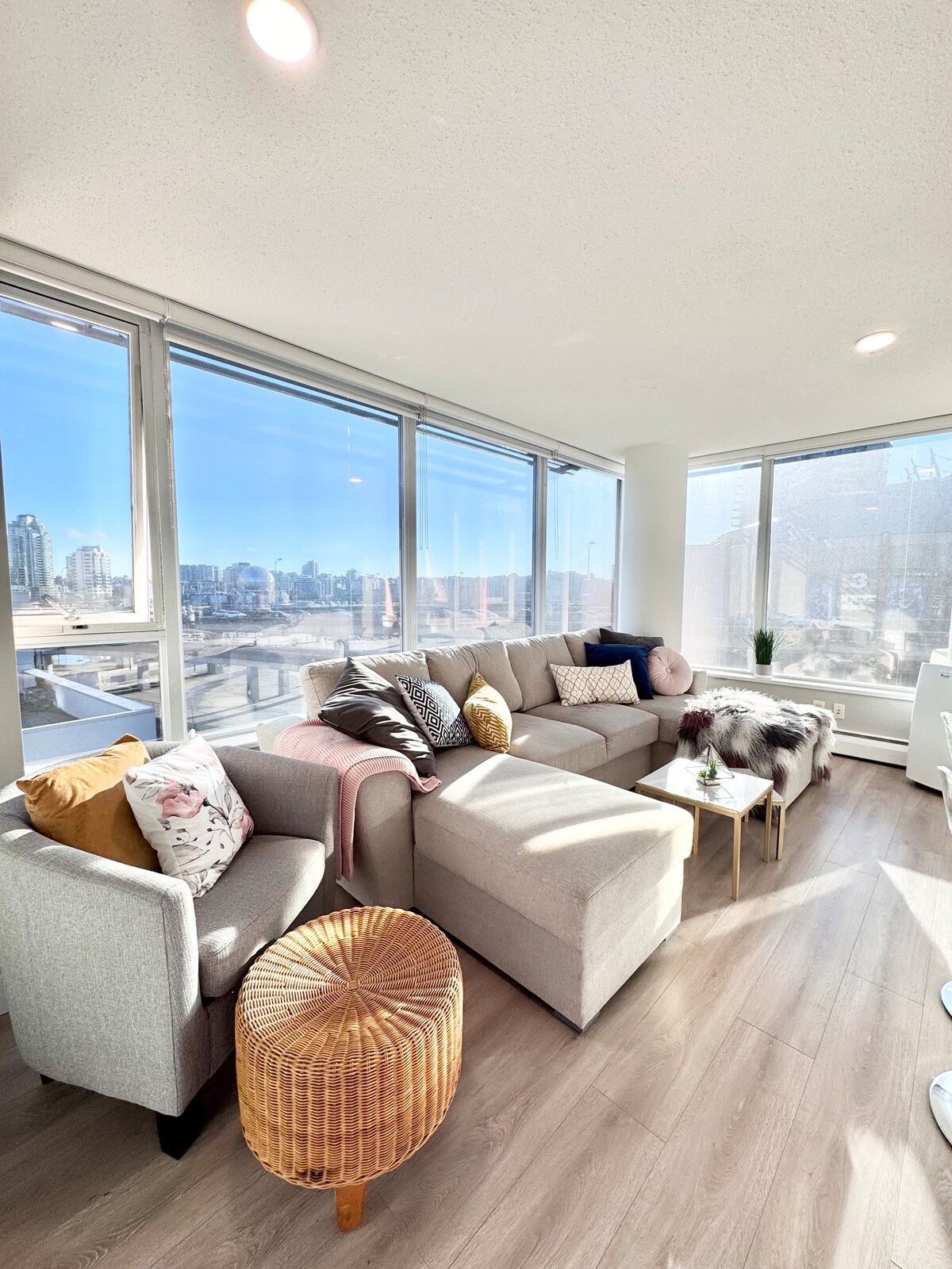 A spacious living area is illuminated by large windows, offering city views. A light-colored sectional sofa is accompanied by two armchairs, adorned with decorative pillows in various colors and patterns. A small round table with a woven design is placed near the seating.